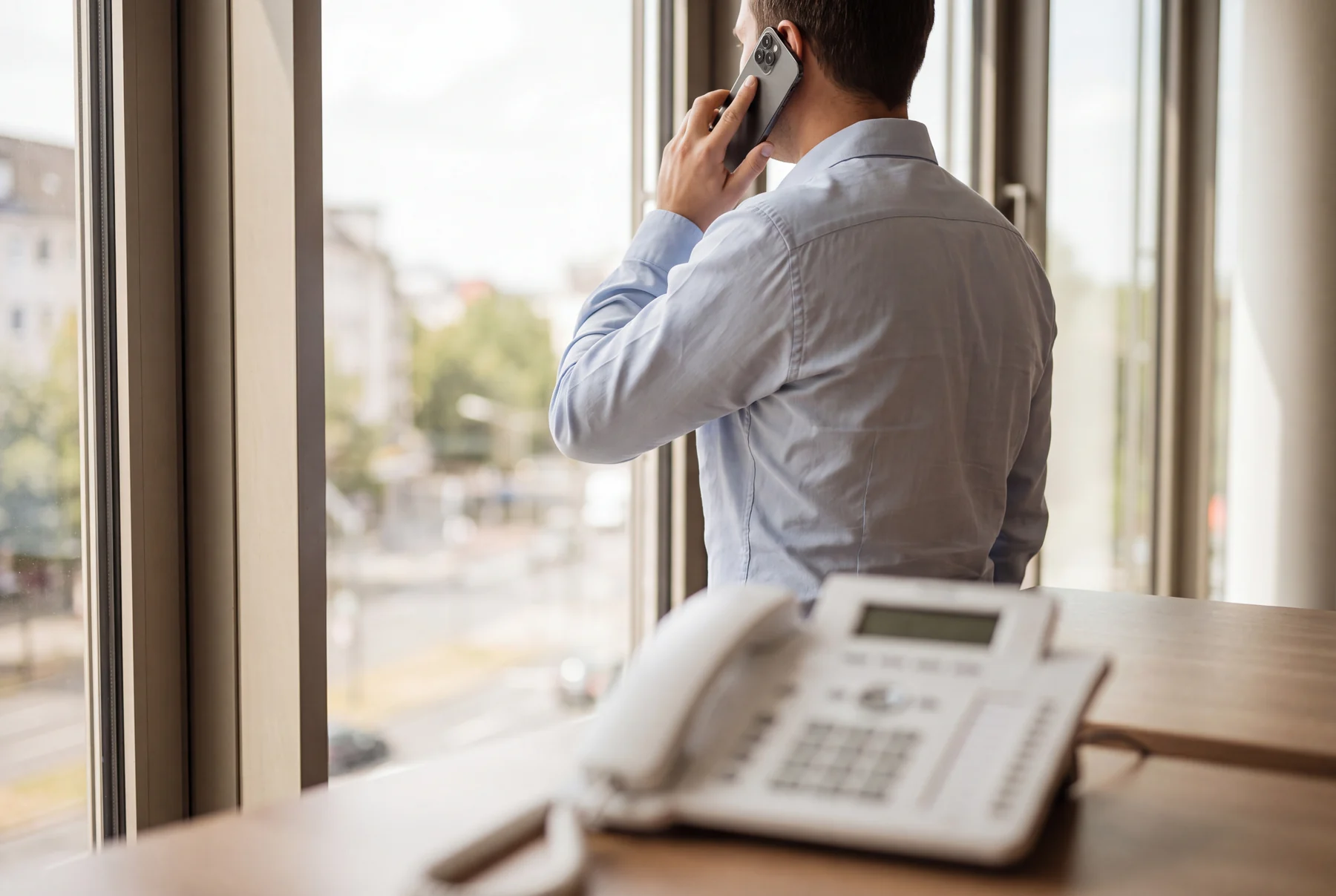 Person am Fenster telefoniert mit dem Handy, im Vordergrund steht ein Tischtelefon auf dem Schreibtisch