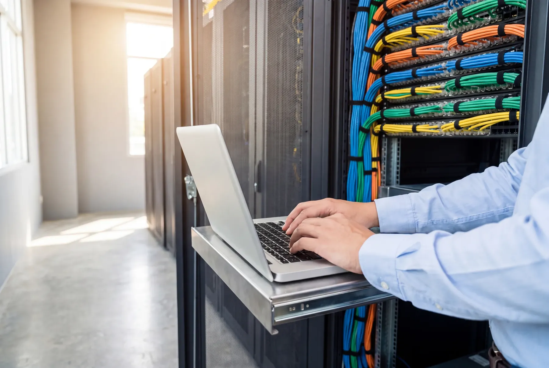 Hände tippen an einem Laptop auf der Auszugsablage eines Netzwerkschranks mit farbig sortierter Verkabelung